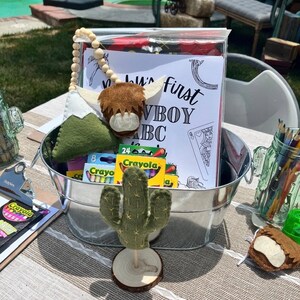 May include: A metal bucket filled with crayons, a felt cactus, a felt highland cow, and a colouring page that says "Baby's First Cowboy ABC".