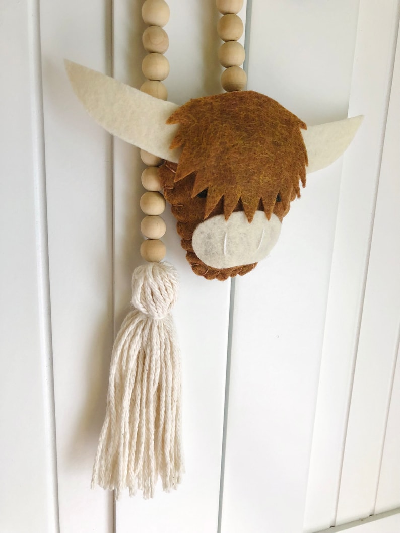 May include: A brown felt Highland cow head with white felt ears and a white felt nose. The cow head is hanging from a wooden bead garland with a white cotton tassel.