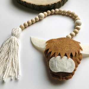 May include: A brown felt Highland cow head with white felt ears and a white felt nose. The cow head is attached to a wooden bead garland with a white tassel.