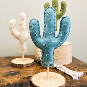 May include: Three felt cactus decorations on wooden bases. The cacti are in shades of green, blue, and white with white stitching. The bases are natural wood.