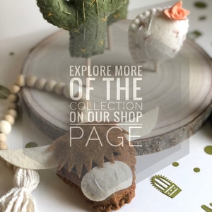 May include: Felt animal figurines, including a green cactus, a white sheep with a pink flower, and a brown highland cow. The figurines are displayed on a wooden slice with a white beaded garland. The text "Explore More Of The Collection On Our Shop Page" is superimposed on the image.