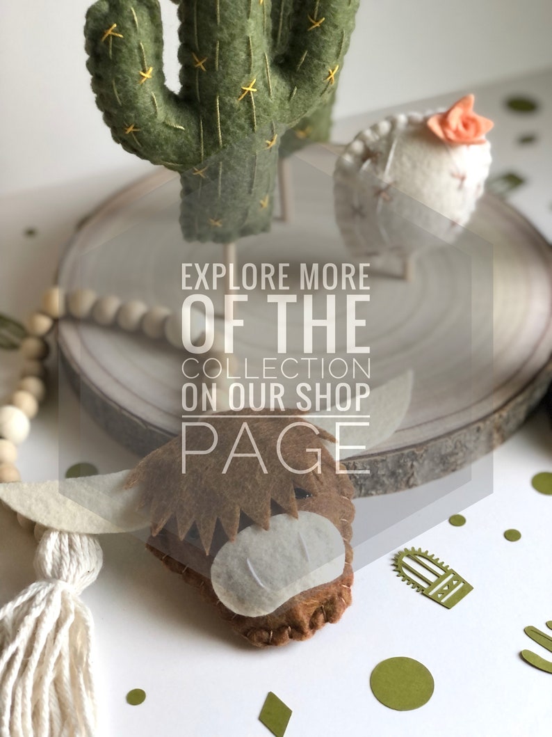 May include: Felt animal figurines, including a green cactus, a white sheep with a pink flower, and a brown highland cow. The figurines are displayed on a wooden slice with a white beaded garland. The text "Explore More Of The Collection On Our Shop Page" is superimposed on the image.