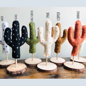 May include: Seven felt cactus figurines in various colours, including white, black, sage green, pink, copper, orange, and cream. Each cactus is standing on a small wooden base.