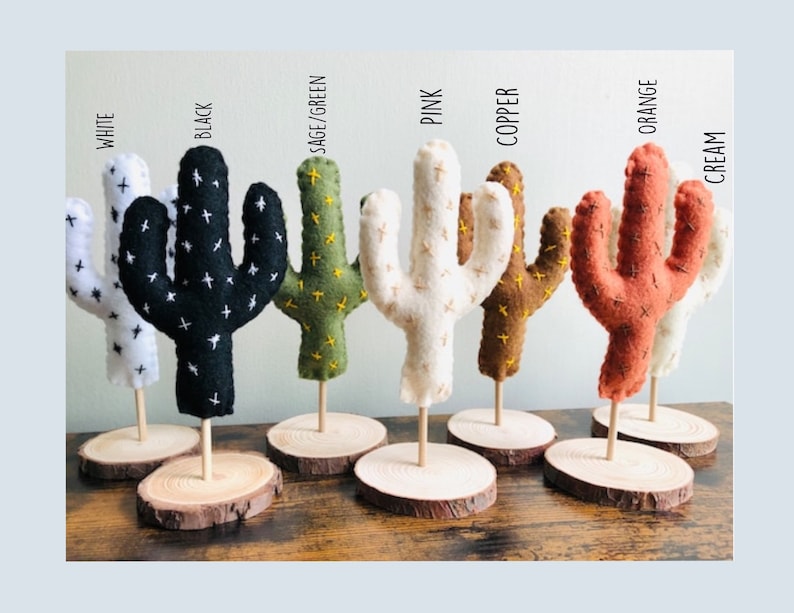 May include: Seven felt cactus figurines in various colours, including white, black, sage green, pink, copper, orange, and cream. Each cactus is standing on a small wooden base.
