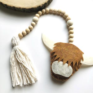 May include: A brown felt Highland cow head with white felt horns and a white felt nose, hanging from a wooden bead garland with a white tassel.
