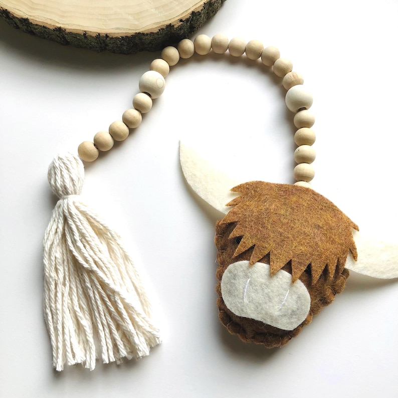May include: A brown felt Highland cow head with white felt horns and a white felt nose, hanging from a wooden bead garland with a white tassel.