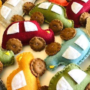 May include: A collection of felt toy cars in various colors, including red, green, blue, yellow, and gray. Each car has white felt windows and brown felt wheels with a stitched cross pattern.