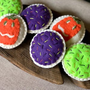 Op de afbeelding: Een verzameling kleurrijk vilten voedsel speelgoed, waaronder groene, paarse en oranje sushi-achtige vormen met geborduurde details. Het speelgoed is gerangschikt op een houten dienblad, wat een speelse en handgemaakte esthetiek laat zien.