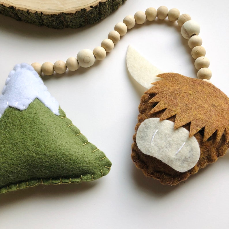 May include: A felt garland featuring a green mountain, a brown highland cow, and wooden beads. The garland is ideal for a nursery or child's room.