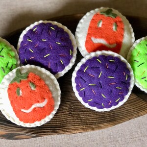 Op de afbeelding: Zes vilten Halloween-cupcakes met verschillende toppings. De cupcakes zijn versierd met groene, paarse en oranje frosting en sprinkles.
