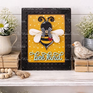 May include: A rectangular wall sign with a black scalloped frame featuring a yellow background with white polka dots. A central bee cutout has white wings, a black and white gingham bow, and a small red heart. The text bee kind is in blue.