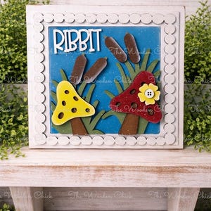 May include: A framed wooden art piece with the word "RIBBIT" in white letters. The artwork features a blue background with wooden cutouts of mushrooms, cattails, and grass. A yellow mushroom with black spots and a red mushroom with a yellow flower are visible.