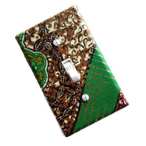 May include: A light switch cover with a green, brown, and gold patterned fabric design. The switch cover has a white toggle switch and two silver screws.
