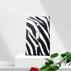 May include: A white light switch cover with a black and white zebra print design. The switch is in the off position. A single red rose is in the foreground.