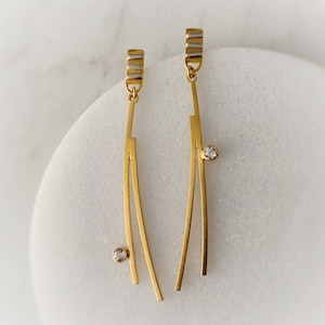 May include: A pair of gold earrings with a single clear stone on each earring. The earrings are made of two thin gold bars that are connected at the top and bottom. The bars are slightly curved.