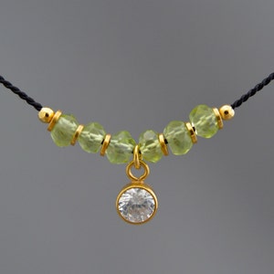 May include: A delicate necklace with a small, clear crystal pendant. The necklace is made of black cord and features a row of light green beads.