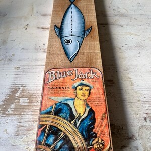 Peut inclure: Une étiquette de boîte de sardines vintage de la marque Blue Jack avec un marin au gouvernail d'un volant de navire. L'étiquette est sur un fond en bois avec un poisson peint bleu et blanc au-dessus.