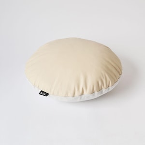 Peut inclure: Un coussin rond en cuir beige avec une surface lisse et légèrement texturée. L'oreiller présente un dessous en tissu blanc et une petite étiquette rectangulaire noire sur le côté avec le texte blanc ni.ni. imprimé en police sans-serif minuscule.