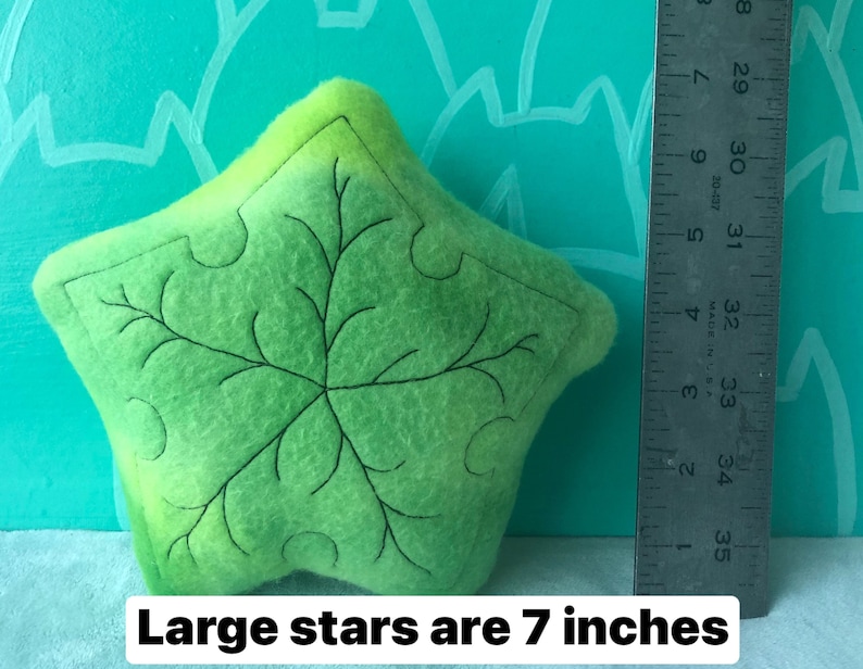 May include: A lime green star-shaped felt pillow with a black embroidered leaf design. The text "Large stars are 7 inches" is at the bottom of the image. The pillow is approximately 7 inches in size.
