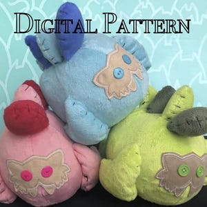 Peut inclure: Trois peluches en forme de hiboux. Les hiboux sont rose, bleu et vert. Chaque hibou a un visage en feutre marron avec deux gros boutons roses pour les yeux. Les hiboux sont disposés en pile, le hibou bleu en haut.