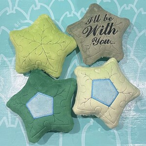 May include: Four star-shaped plush pillows in shades of green and beige. Two pillows have a leaf design, one has a blue pentagon, and another has the text "I'll be With You...". The pillows are on a light blue surface.