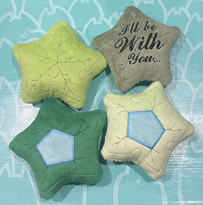 May include: Four star-shaped plush pillows in shades of green and beige. Two pillows have a leaf design, one has a blue pentagon, and another has the text "I'll be With You...". The pillows are on a light blue surface.