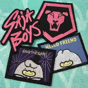May include: A collection of embroidered patches with various designs. One patch features the text "SIX BOYS" in pink on a black background. Another shows a pink lion's head on a black pentagon. Two other patches have cartoon characters and text.