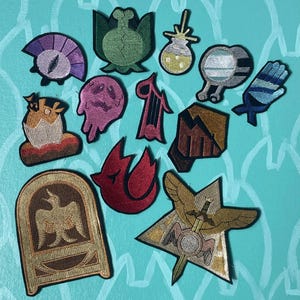 May include: A collection of 12 embroidered iron-on patches featuring various designs, including a bird, a star, a sword, a mountain, a ghost, a plant, a potion, a bird in a cage, a fire, an egg, a hand, and an eye. The patches are colorful and detailed, and would be perfect for adding a unique touch to clothing, bags, or other items.
