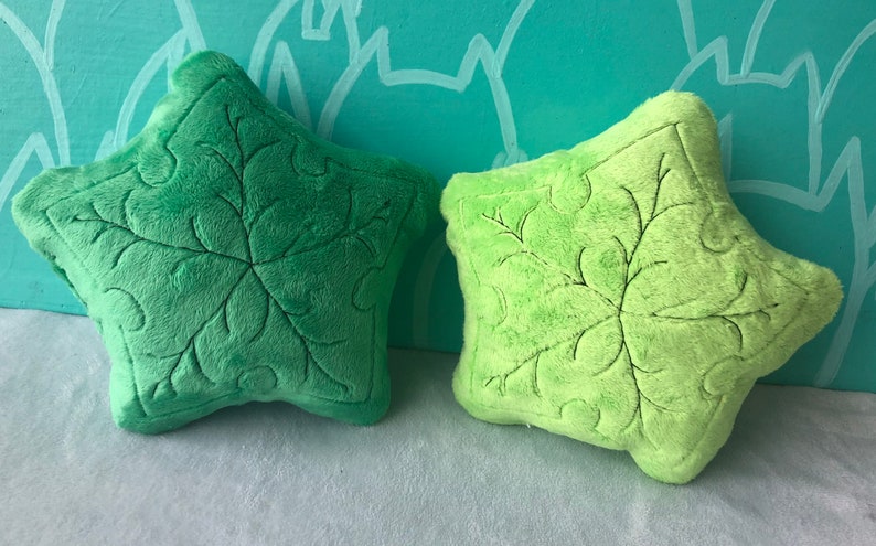May include: Two plush leaf-shaped pillows in shades of green. The pillows have detailed embroidered vein patterns and are set against a light blue background with a leaf pattern. The pillows are soft and fluffy.