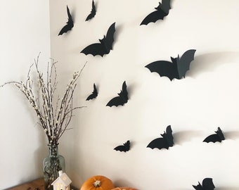 May include: A Halloween-themed display featuring black paper bats affixed to a white wall. The scene includes orange pumpkins of varying sizes, a glass bottle with branches, and a small white house-shaped candle holder, creating a festive autumn ambiance.