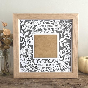 May include: A square light wood frame containing a white paper cutout with a botanical pattern of leaves and pinecones on a gray background. The center features a square opening with the text JAMES & HAYLEY and ENGAGED 13.09.24.
