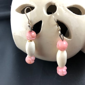 Può includere: Un paio di orecchini pendenti con perline rosa e bianche. Gli orecchini sono appesi a un supporto in ceramica bianca con tagli.