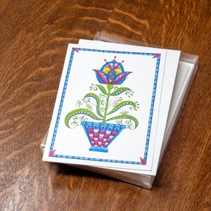 May include: A stack of blank greeting cards with a colorful illustration of a stylized flower in a pot on the front. The flower is blue, pink, yellow, and green. The pot is blue, pink, and white.