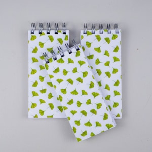 May include: Three spiral bound notebooks with a white background and a repeating pattern of green ginkgo leaves.