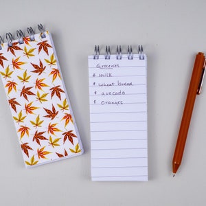 Puede incluir: Un bloc de notas blanco con una lista de la compra escrita a mano que dice "Groceries: milk, wheat bread, avocado, oranges". También se ve una pluma marrón y un pequeño bloc de notas con un patrón de hojas de otoño.