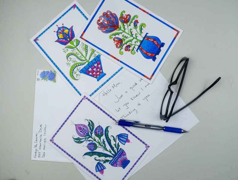 May include: Four hand-drawn postcards with colorful floral designs in blue, red, purple, and green. The postcards are on a white surface with a pen, glasses, and an envelope.
