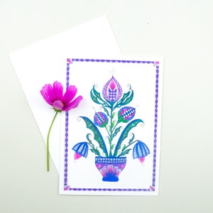 May include: A hand-drawn illustration of a stylized floral design with blue, green, and pink flowers in a blue and white patterned pot. The illustration is framed with a purple border.