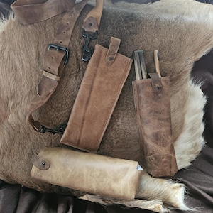 May include: A collection of handcrafted leather knife sheaths and a shoulder strap in various shades of brown. The sheaths are designed to hold knives, and the strap features a metal buckle and clasp. The items are arranged on a textured, light brown surface.