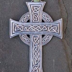 Può includere: Un ornamento a forma di croce celtica in argento con intricati disegni di nodi. La croce è sospesa da un nastro verde.