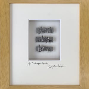 May include: A framed print with the words "faith, hope, love" in a silver font. The words are arranged vertically and are framed in a wooden frame. The print is signed "Cynthia Webb" in cursive.