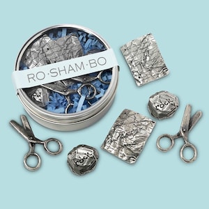May include: A silver-colored tin with the text "RO SHAM BO" on a white band. Inside the tin are scissors, paper, and blue paper shreds. Scattered around the tin are two pairs of scissors, two rock-shaped objects, and two paper-shaped objects.