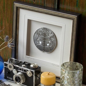 May include: A framed silver-colored relief sculpture with a tree and two doves, labeled "perfect union." A vintage black camera and a yellow candle sit nearby. The frame is silver with a white mat.