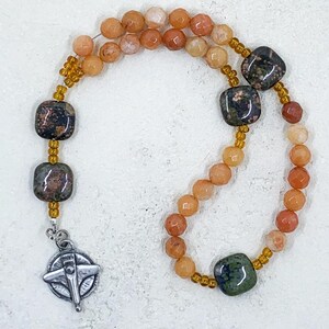 May include: A prayer bead necklace with a silver airplane charm. The beads are made of orange, brown, and green stones.