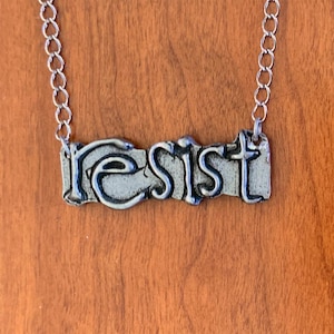Könnte beinhalten: Eine silberne Halskette mit einem Anhänger, auf dem in einer stilisierten Schrift "resist" steht.