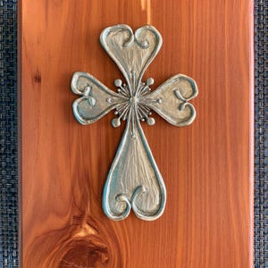 May include: A silver-colored cross with heart-shaped ends is mounted on a rectangular, reddish-brown wooden plaque. The cross has a detailed, ornate design with a central starburst. The wood grain is visible, adding texture to the piece.
