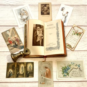 May include: An open book titled "Missel Romain" with a sepia-toned image of a woman and child, surrounded by vintage religious cards and a brass object. The cards feature religious figures and scenes, with various designs and colours.