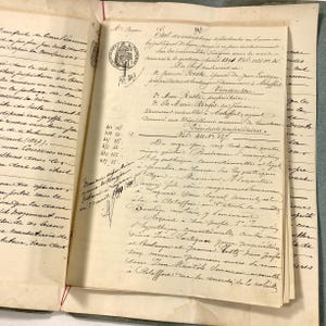 May include: An antique French legal document with handwritten cursive text in black ink on off-white paper. The document features a circular stamp with the text Republique Francaise and lists names such as Jeanne Reste and Marc Reste. The pages are aged with visible yellowing and are bound together with thin red thread.