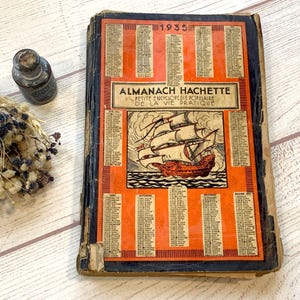 Puede incluir: Libro vintage "Almanach Hachette" de 1935 con portada naranja y una ilustración de barco. El libro tiene lomo negro y muestra signos de desgaste. Una pequeña botella de vidrio y flores secas están junto al libro.