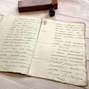 May include: An open antique book with handwritten script on aged paper. A small, dark brown ink bottle and a wooden box with the word "Format" are on the surface. The book's pages are yellowed with age.