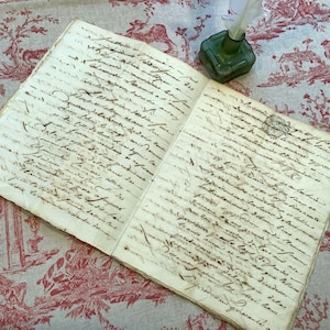 Puede incluir: Un libro antiguo abierto con escritura manuscrita en tinta marrón. Un pequeño tintero de vidrio verde con una pluma blanca se encuentra a la derecha. El libro y el tintero descansan sobre una tela estampada en rojo y blanco.